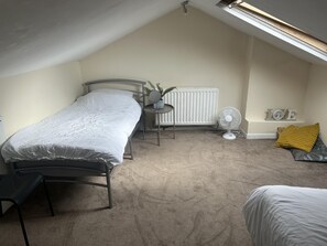 Quarto com cama, uma mesinha com uma planta, radiador, ventilador e janela com vista para o céu.