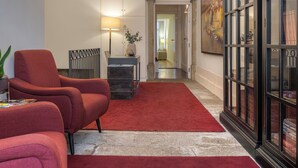 Un couloir avec un tapis rouge, un lustre, un fauteuil rouge, une lampe et un grand tableau accroché au mur.