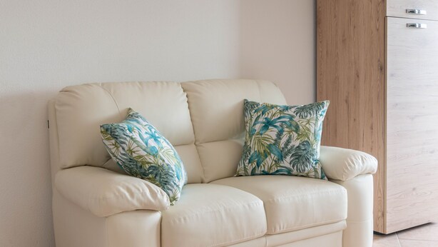 Un canapé en cuir beige, agrémenté de deux coussins décoratifs, est placé devant un mur uni, près d’un meuble en bois.