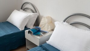 Un lit simple avec un cadre en métal, des oreillers blancs, une couvre-lit bleue et une table de chevet sur laquelle se trouvent un magazine et un oreiller blanc.