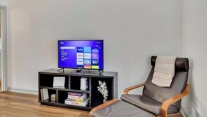 A living room with a flat-screen TV on a stand, a leather chair, and a small shelf with books and decorative items.