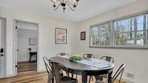 A dining room with a round table, chairs, a chandelier, and a window with a view of trees.
