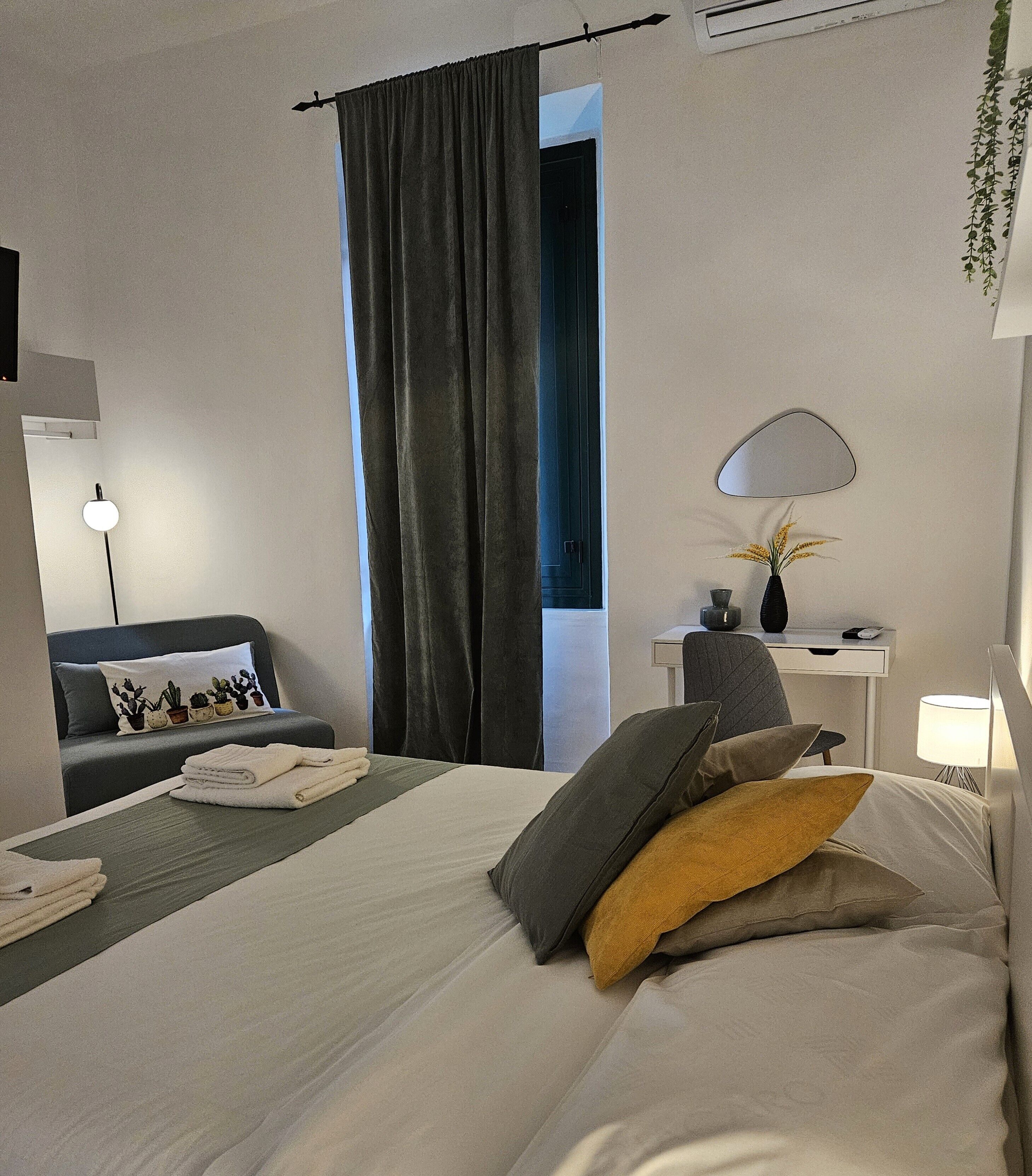 A hotel room with a bed, a desk, a chair, a lamp, a mirror, and a window with curtains.