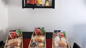 Un coin repas avec une étagère noire où sont posés des bocaux en verre, une plante en pot et un pot de miel. En dessous, on trouve trois boîtes en bois contenant du pain et des viennoiseries.