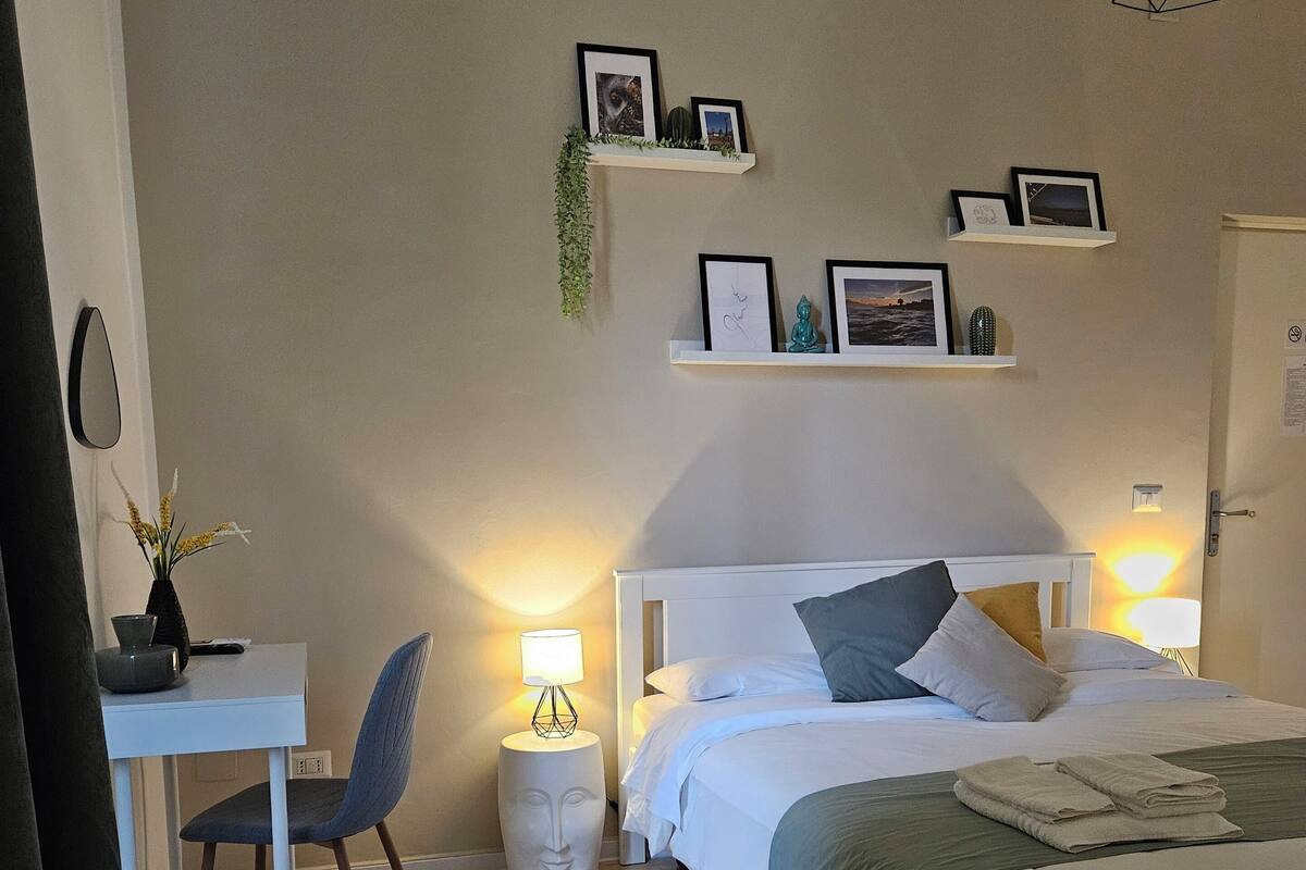 A bedroom with a bed, bedside tables, a desk, and a chair. There are wall-mounted shelves with pictures and a hanging light fixture.