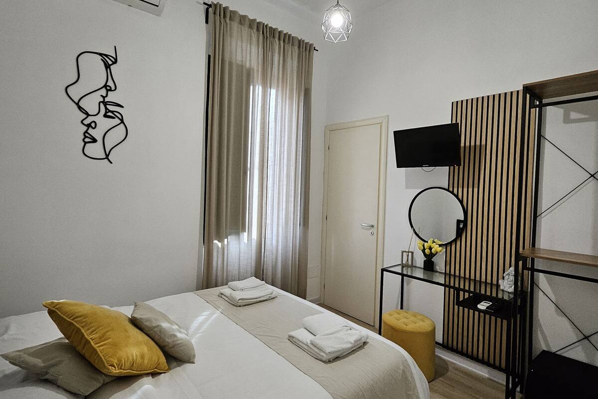 A modern bedroom with a large bed, a bedside table with a mirror, and a TV.