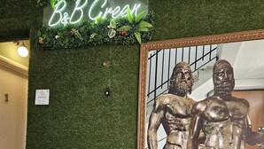 Un miroir reflétant deux statues de bronze, une enseigne au néon indiquant « B&B Green », et un mur vert avec une petite pancarte indiquant « VERT ».