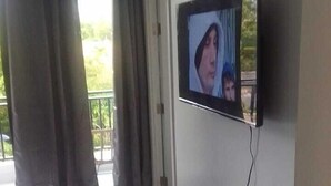Una habitación con una televisión de pantalla plana en la pared, una puerta y vistas a un balcón con cortinas.