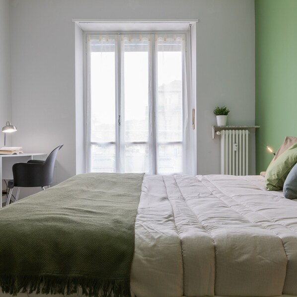 A bedroom with a bed, a desk, a chair, a radiator, and a window with curtains.