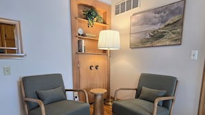 Una habitación con dos sillones tapizados grises, un aparador de madera con una planta, una lámpara y un cuadro de un paisaje.