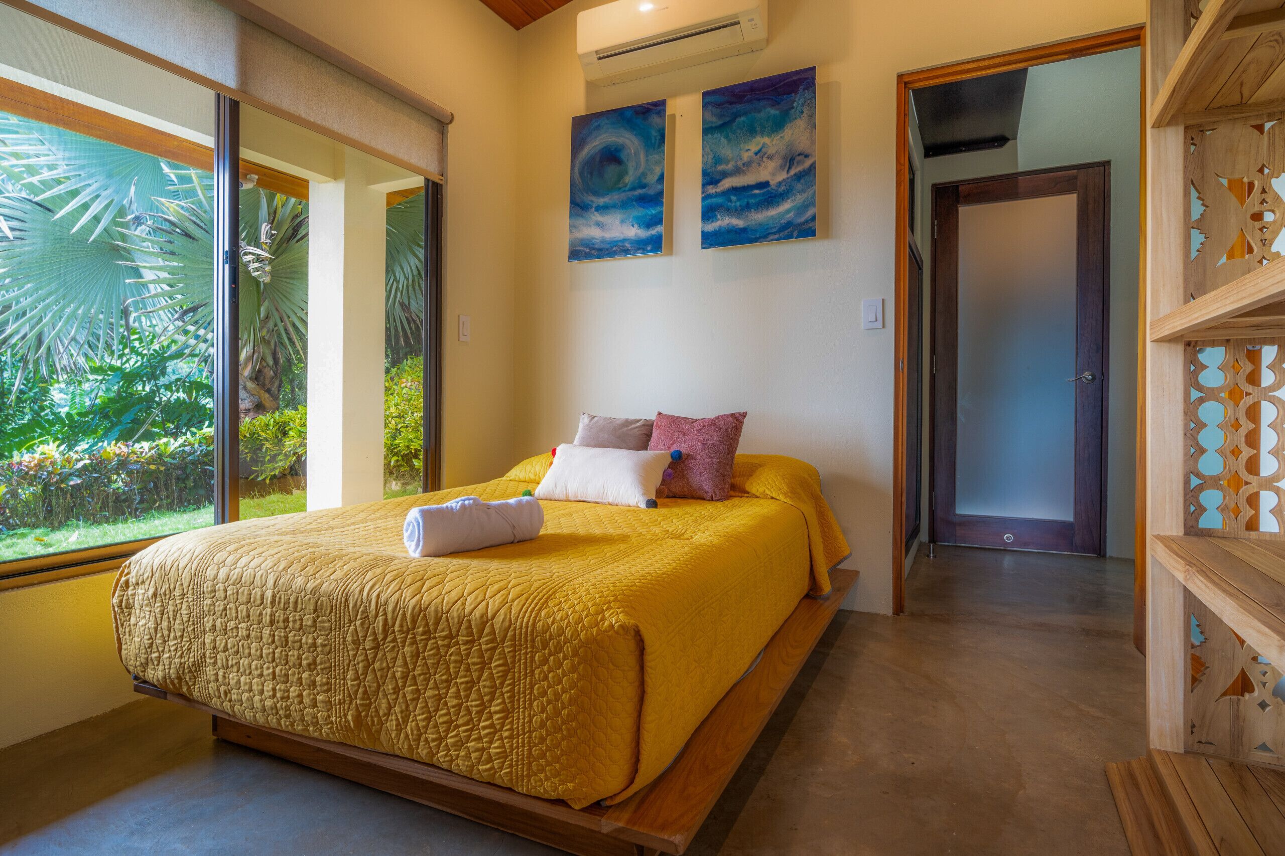 A bedroom with a large bed, a window with a view of greenery, and two paintings on the wall.