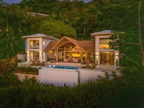 A modern two-story house with a swimming pool, surrounded by lush greenery and a thatched-roof pavilion.