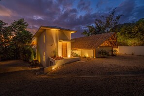 Ein modernes Haus mit einem strohgedeckten Anbau, einem gut beleuchteten Eingangsbereich und einer Auffahrt mit Kies.