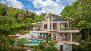Una casa de varios pisos con piscina, rodeada de abundante vegetación.