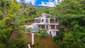 Una casa moderna de dos pisos con piscina, rodeada de exuberante vegetación y un paisaje montañoso.