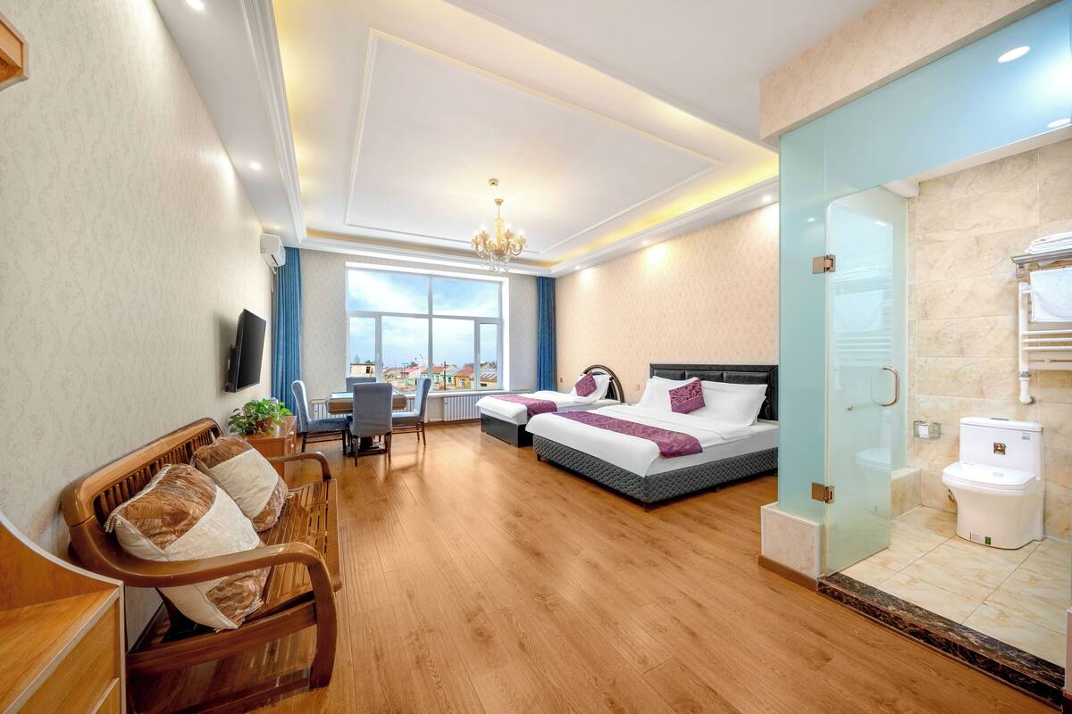Ein Hotelzimmer mit Holzboden, einem Bett mit weißer und violetter Bettwäsche, einem Schreibtisch mit Stuhl, einem an der Wand montierten Fernseher und einem großen Fenster mit Blick auf Gebäude.