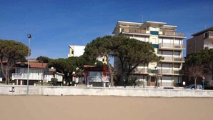 A beachfront property with a sandy beach, a white building with multiple balconies, and a few trees. - Holiday apartment mit Klimaanlage und Balkon (San Michele al Tagliamento)
