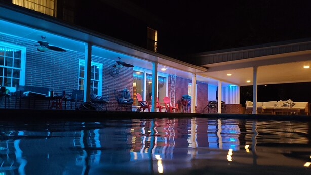 Vista nocturna de un área junto a la piscina con un edificio de ladrillo, mobiliario exterior y un porche cubierto.