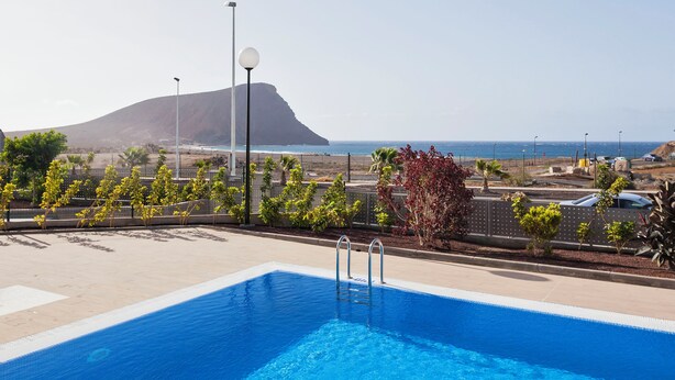 A swimming pool with clear blue water, surrounded by a landscaped area with small trees and shrubs. In the background, there is a view of the ocean and a distant mountain. - Ocean Paradise 1 La Tejita (Intero appartamento, due camere e un bagno) (Granadilla de Abona)
