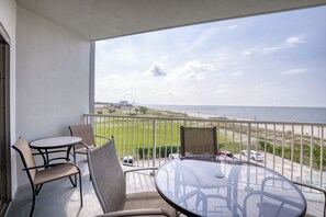 Un balcon avec une table et des chaises en verre, donnant sur une plage délimitée par une clôture et où sont garées des voitures.