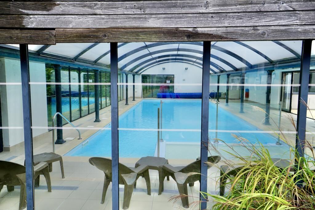 Uma piscina com paredes de vidro transparente e teto de madeira, cercada por cadeiras e plantas em vasos.