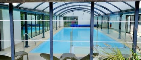 A swimming pool with clear glass walls and a wooden ceiling, surrounded by chairs and potted plants.