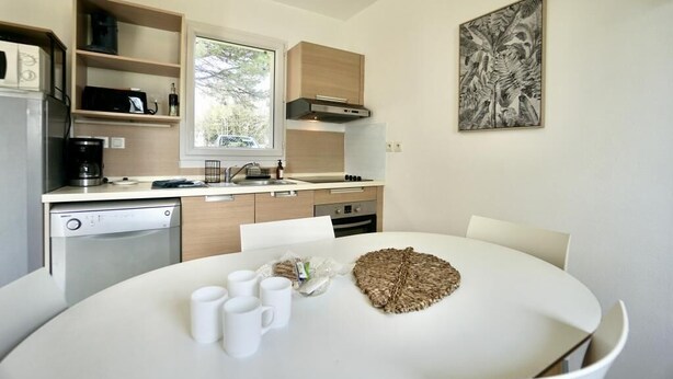 A modern kitchen with a dining area, including a white table with chairs, a coffee machine, and a window with a view of trees.