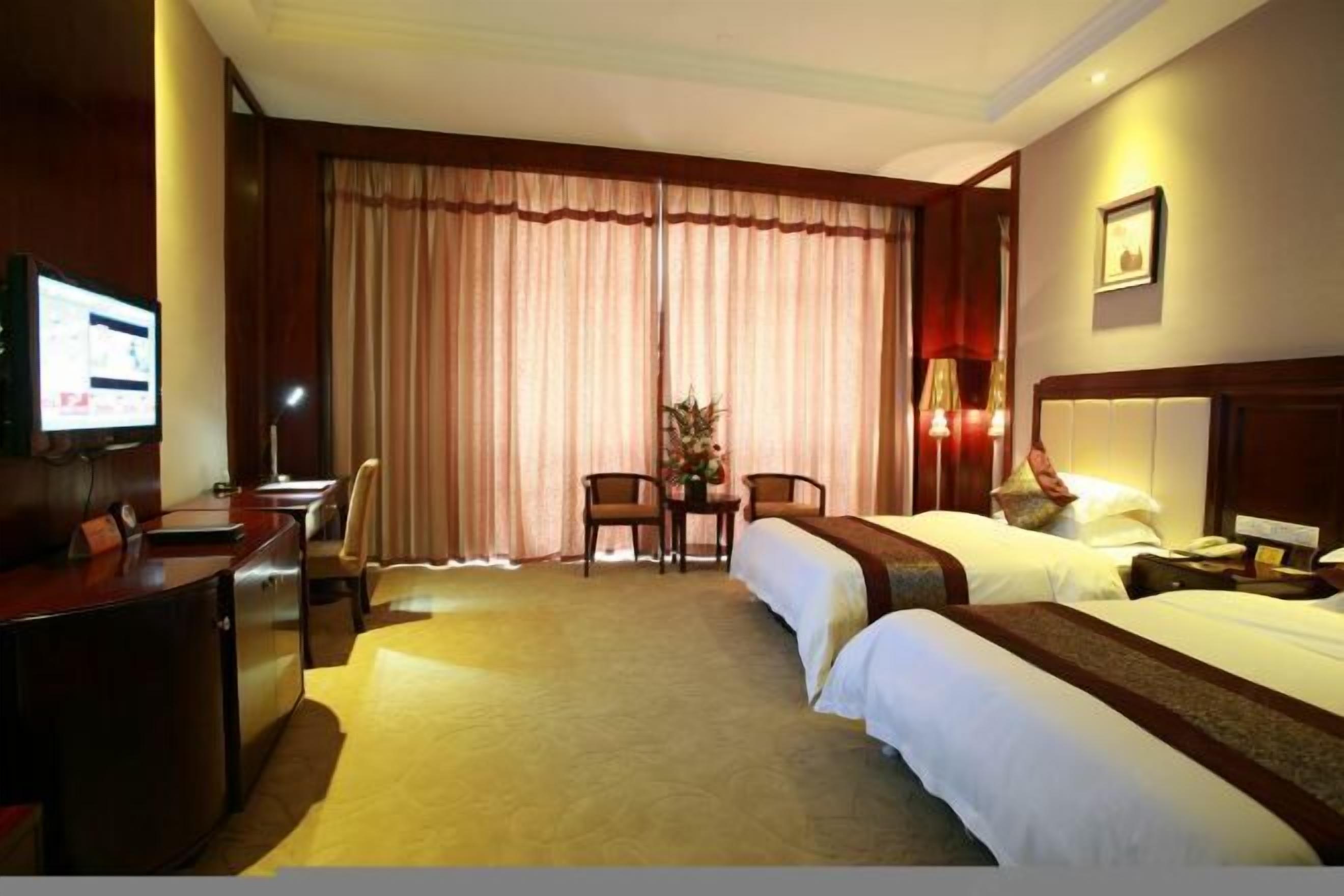 A hotel room with two beds, a desk, a chair, a TV, and a large window with curtains.