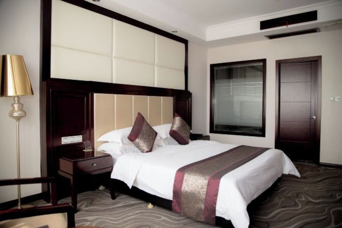 Ein Hotelzimmer mit einem großen Bett, einem Nachttisch, einer Lampe und einem Fenster mit Jalousien.
