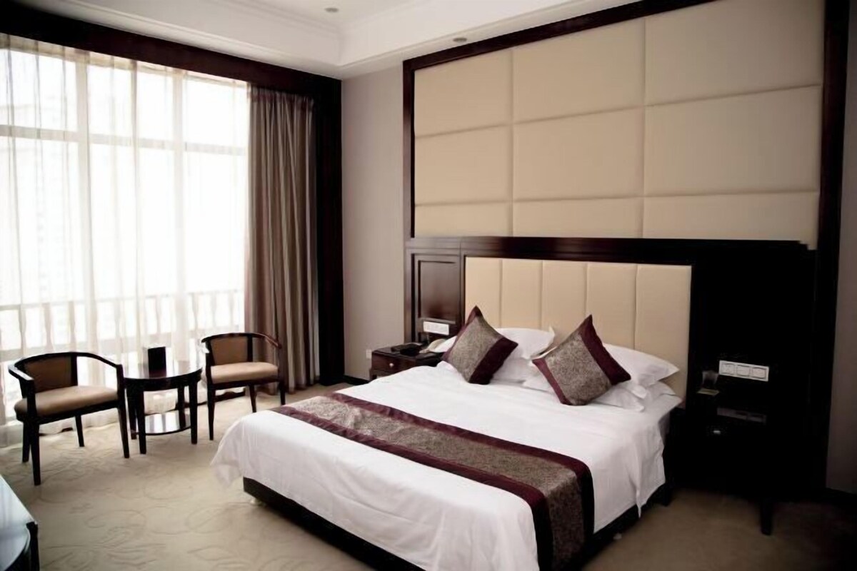 A hotel room with a large bed, two chairs, a nightstand, and a window with curtains.