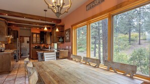 Une cuisine rustique avec des meubles en bois, une grande table à manger et une vue sur la forêt grâce à de grandes fenêtres.