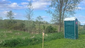 Eine grüne mobile Toilette in einer Wiese mit Baum und blauem Himmel im Hintergrund.