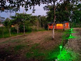 Un paisaje rural con una pequeña estructura naranja, posiblemente un cobertizo o depósito, rodeado de árboles y un camino de tierra.