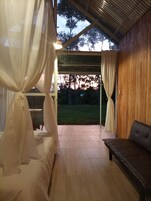 Una habitación con cama, un banco de madera y vista a un área verde a través de una ventana con cortinas.