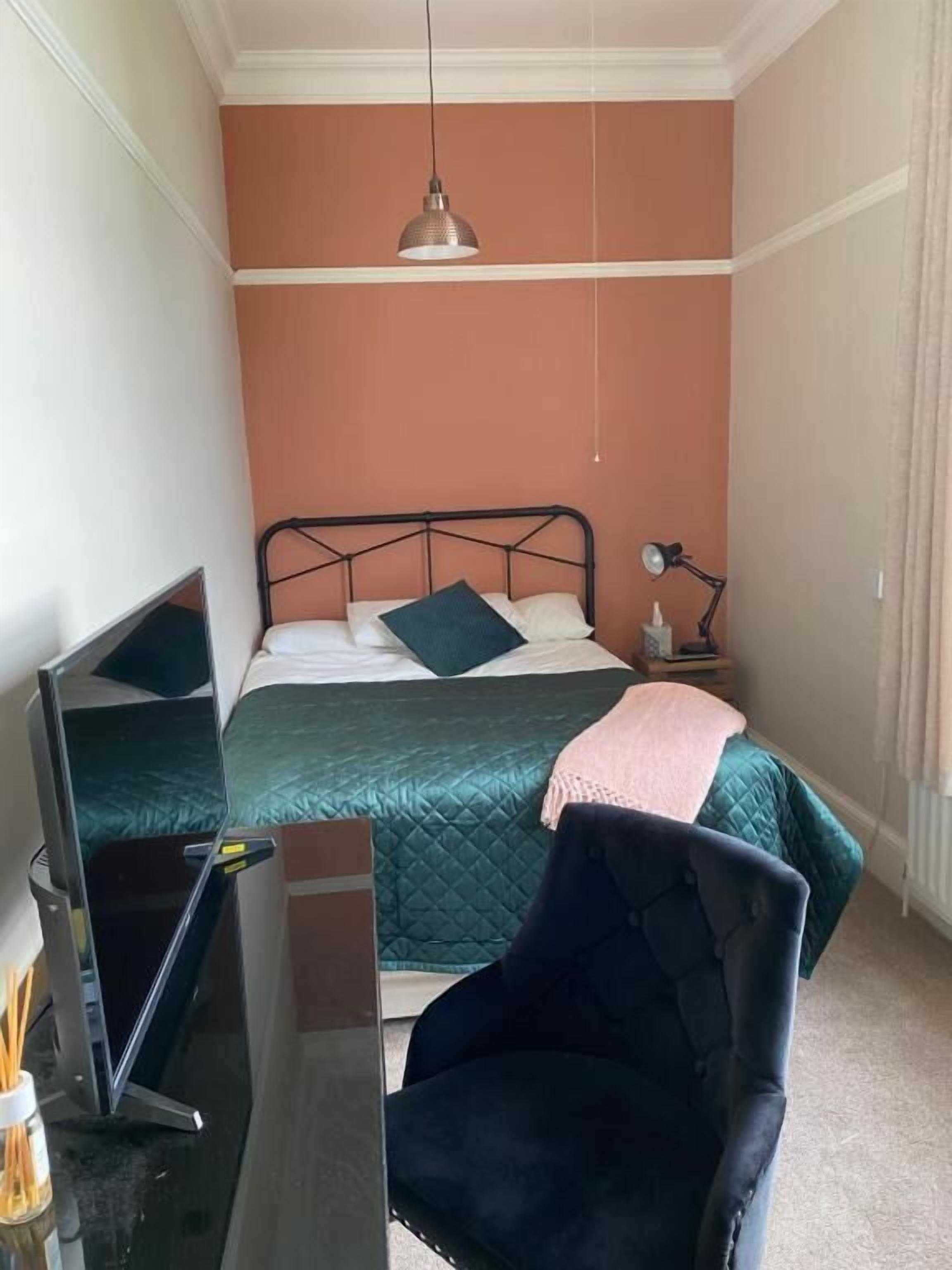 Un dormitorio con cama, mesita de noche, lámpara, silla y escritorio con computadora.