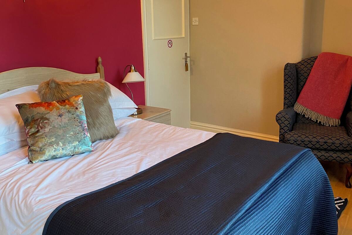 A bedroom with a bed, pillows, a chair, a door, and a wall-mounted light fixture.