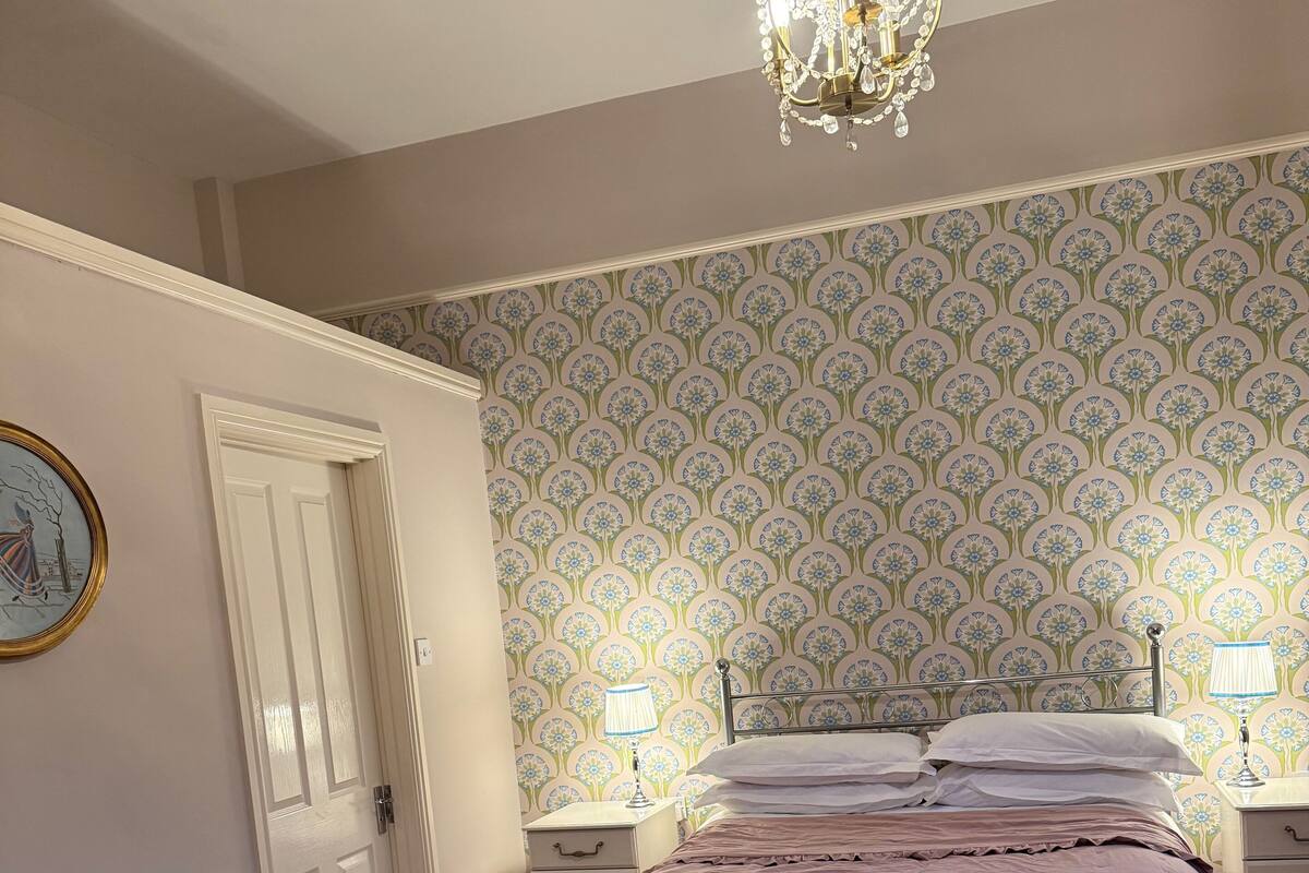 A bedroom with a bed, two bedside tables with lamps, a chandelier, a door, and a patterned wallpaper.