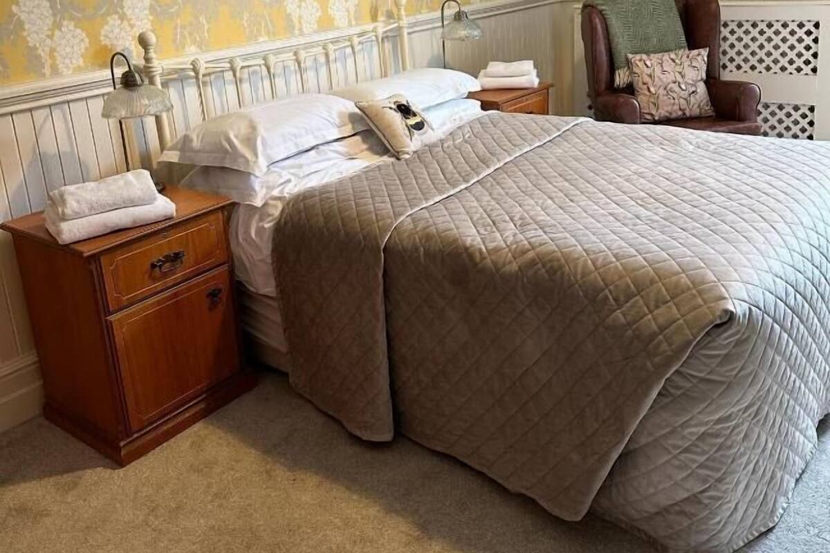 A neatly made bed with a quilted cover, a wooden nightstand with a lamp, and a chair with a blanket.