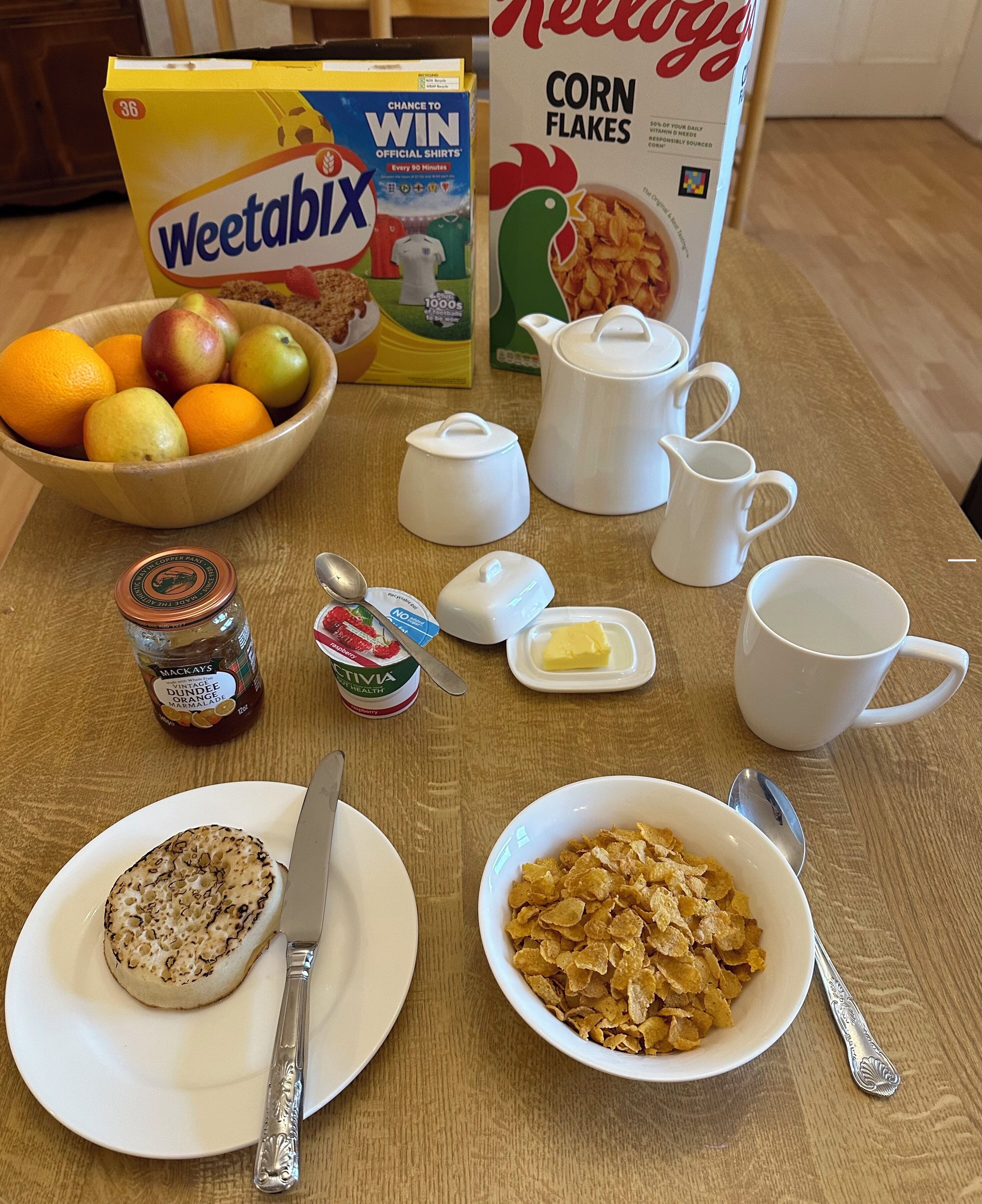 A breakfast setup with cereal, fruit, butter, jam, and a teapot.