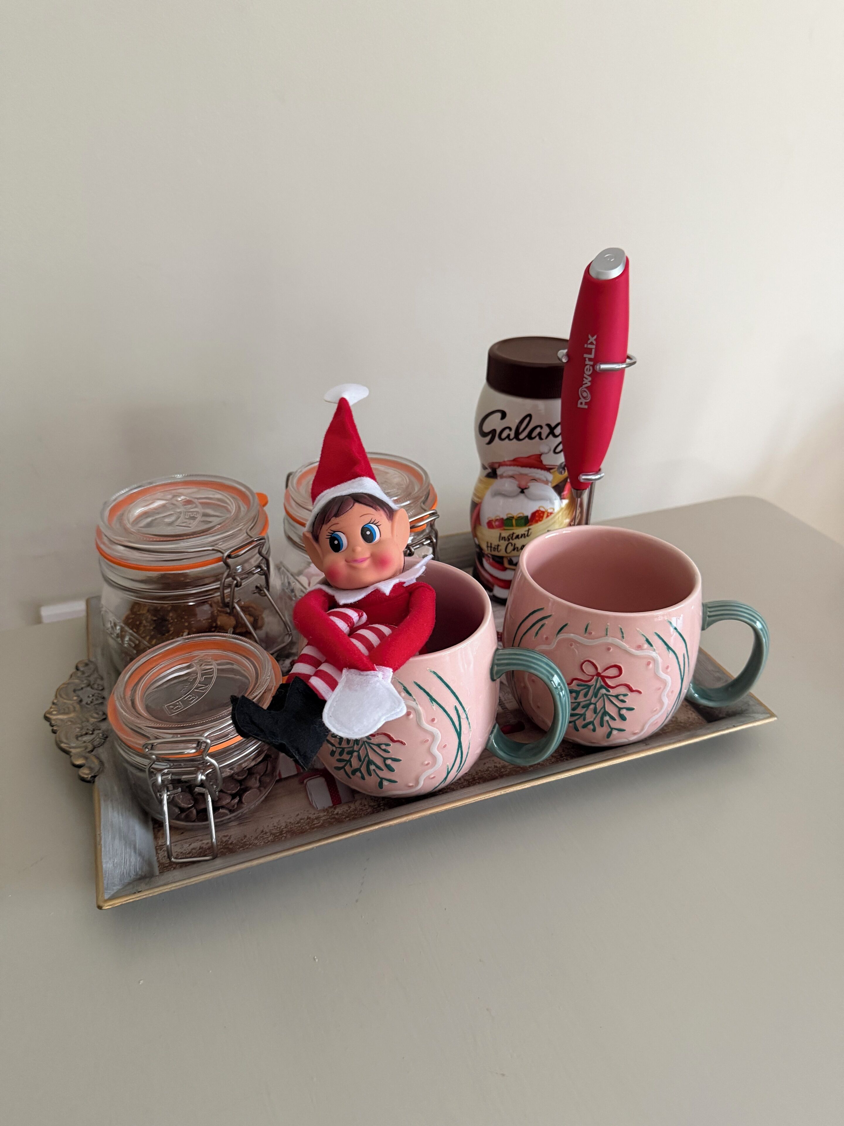 A tray with a decorative elf figurine, two glass jars, a pink mug, and a red-handled utensil.