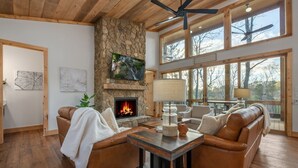 A cozy living room with a stone fireplace, leather sofas, and a large window overlooking a forest. - Enchanted Ridge Retreat: Family friendly, peaceful property, hot tub, sleeps 10 (Ellijay)