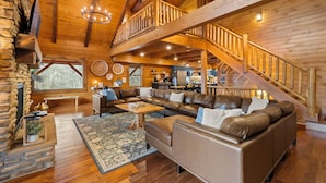 A spacious living area with a large brown leather sofa, a wooden coffee table, and a stone fireplace. A wooden staircase leads to an upper level.