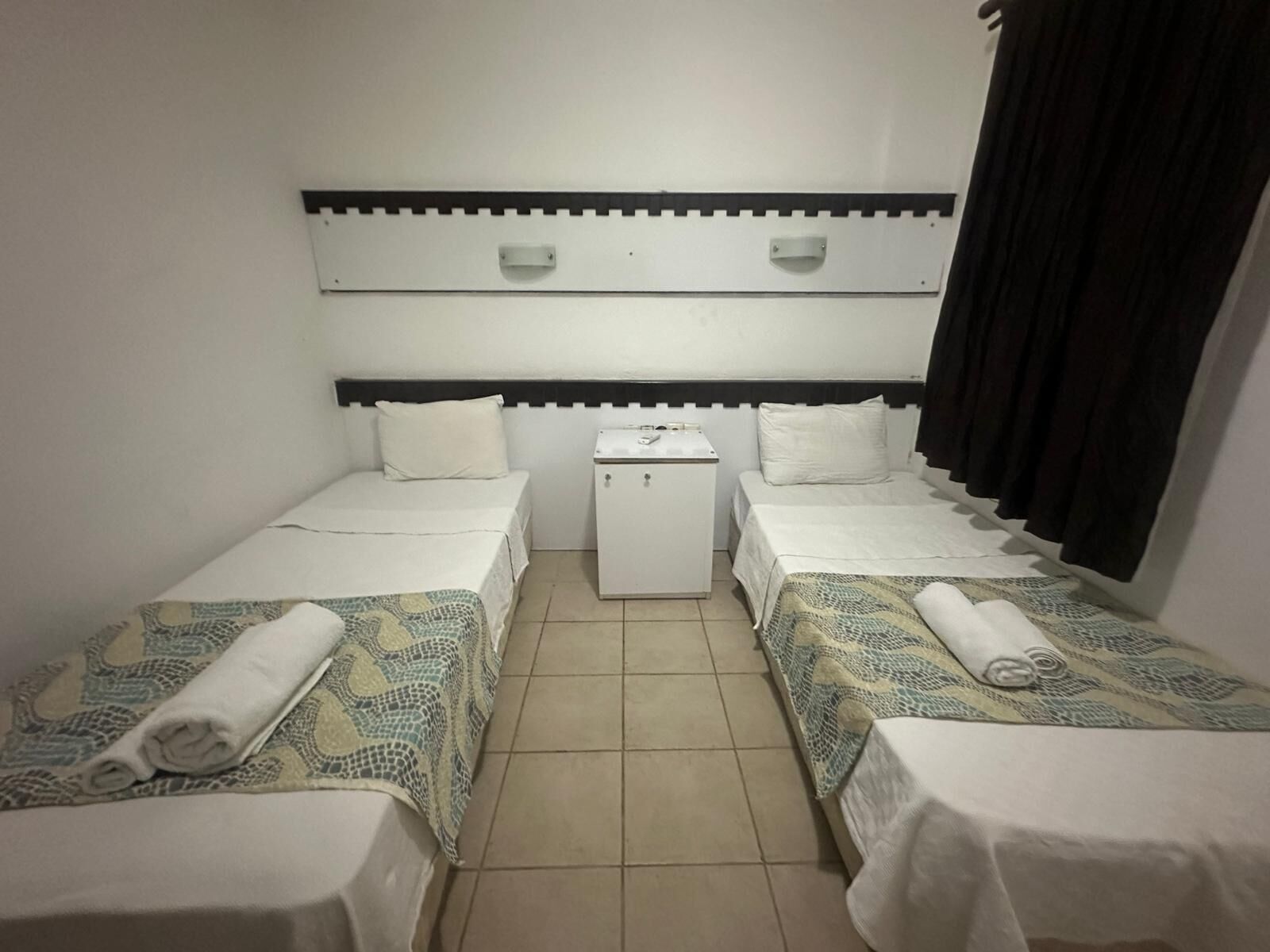 A small, clean room with two beds, a small white cabinet, and a window with black curtains.