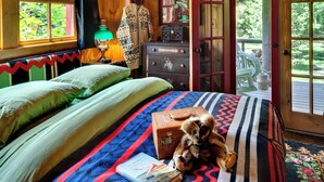 Interior de una cabaña de madera con una cama, un aparador, una lámpara y una puerta que da al exterior.