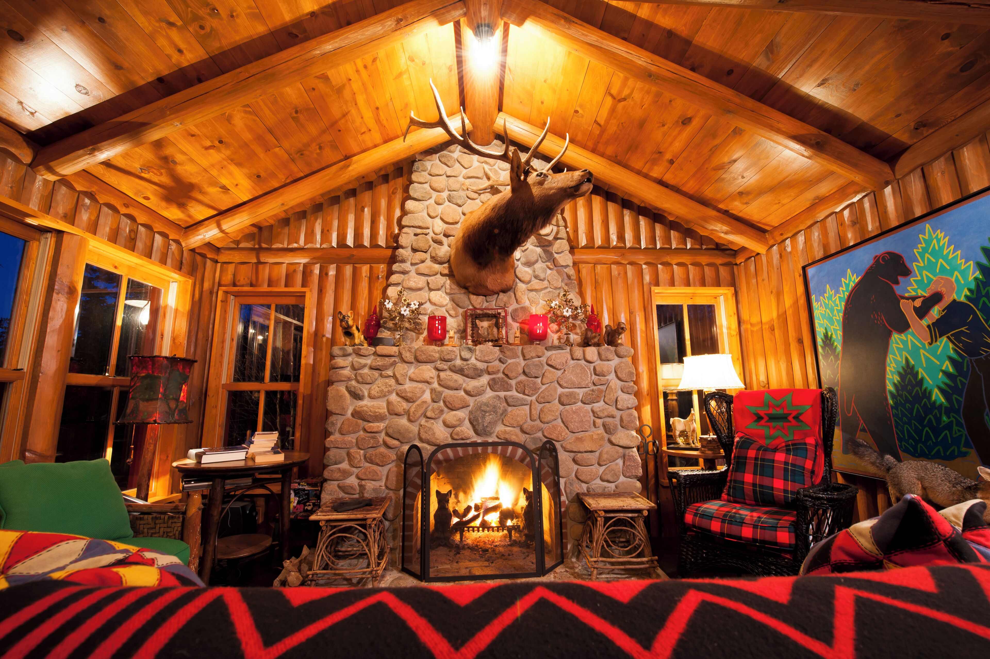 A log cabin interior with a stone fireplace, wooden walls, and a mounted deer head.