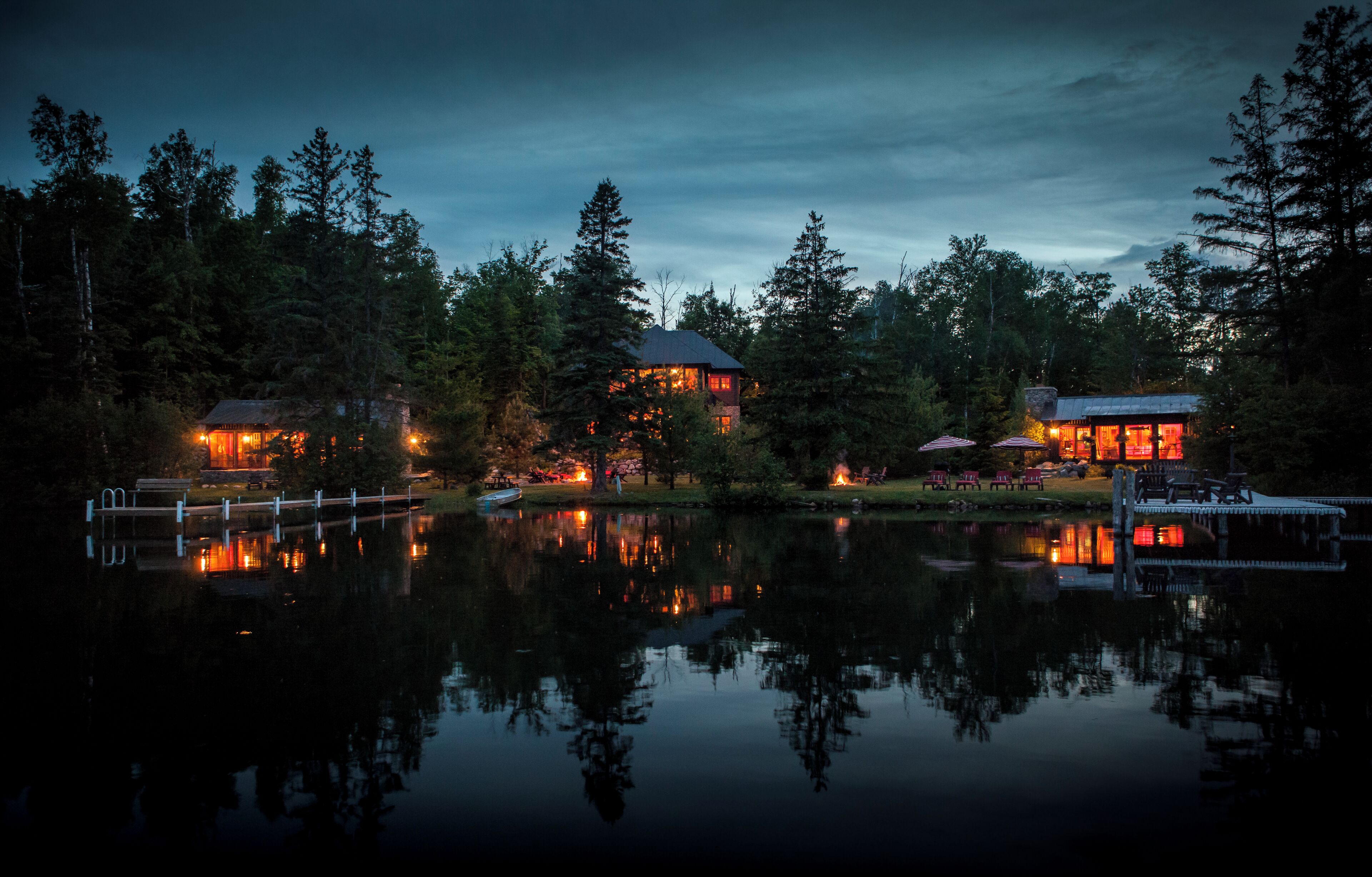 Une propriété au bord du lac, comprenant deux cabanes illuminées, un quai et dont l’image se reflète dans l’eau.