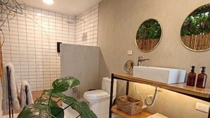 Une salle de bain moderne avec des toilettes blanches, un lavabo avec un miroir, une étagère en bois avec des serviettes et une grande plante verte.