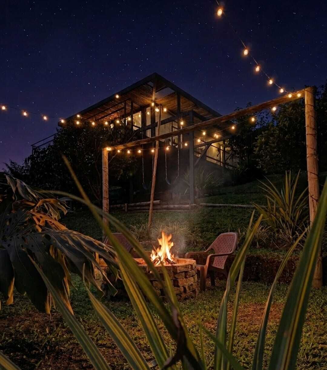 Une structure en bois dotée d’un foyer et illuminée de guirlandes lumineuses, entourée de chaises et de plantes.