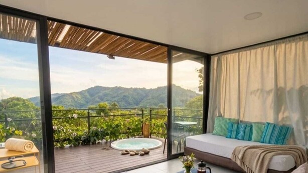 Une chambre moderne dotée d’une grande porte-fenêtre coulissante donnant sur un balcon, d’un canapé, d’une petite table sur laquelle se trouve un plateau de fruits et une cafetière, et offrant une vue sur les montagnes.