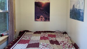 Une chambre avec une couverture à motifs, une affiche représentant un paysage de montagne et une fenêtre avec des rideaux.